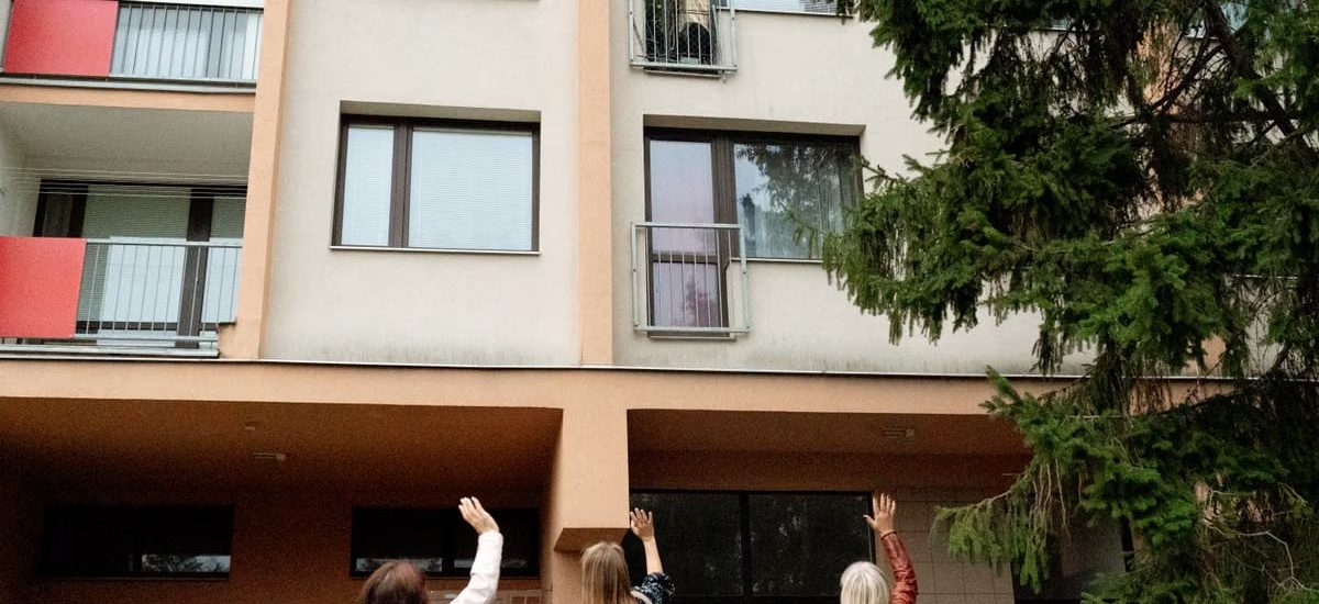 Three women waving to an elderly woman on an apartment balcony in Prague.