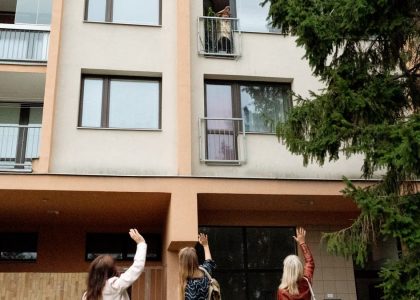 Three women waving to an elderly woman on an apartment balcony in Prague.